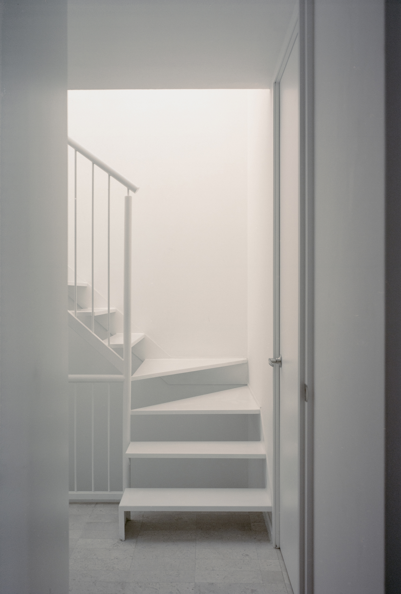 House Within a Cube I by operadora, photographed by Arturo Arrieta — Film photograph of the stair framed by a doorway, treads turning up past a slim railing, a door at right.