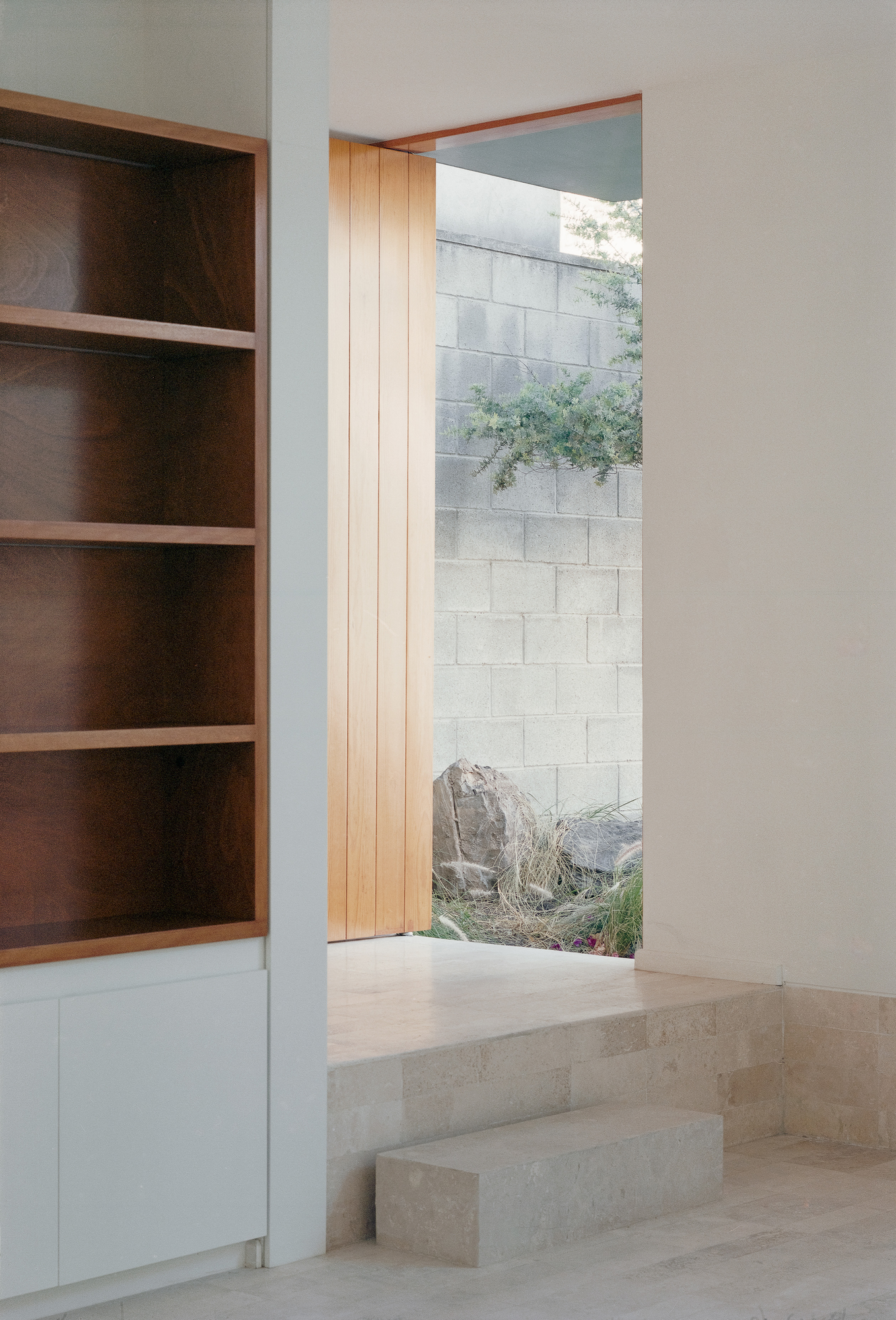 House Within a Cube I by operadora, photographed by Arturo Arrieta — Film photograph of built-in shelving at left, an open door, and steps rising to the threshold, a rock and grasses outside against the wall.
