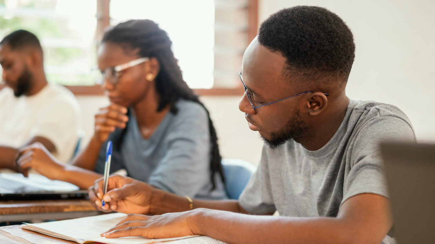 Three students with glasses studying together at a desk with notebooks and laptops.