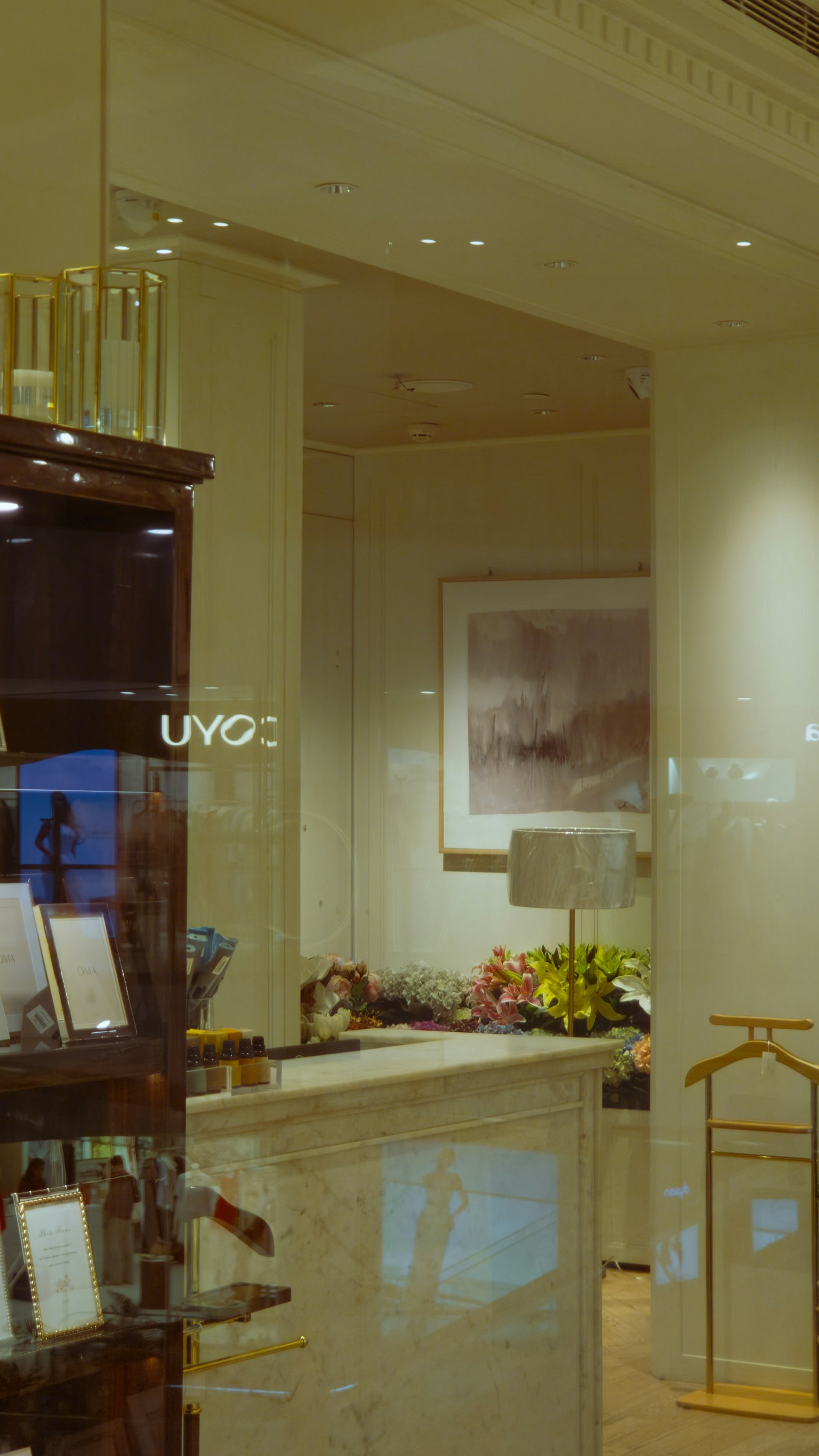 A boutique window at dusk with a framed painting, a lamp and flowers