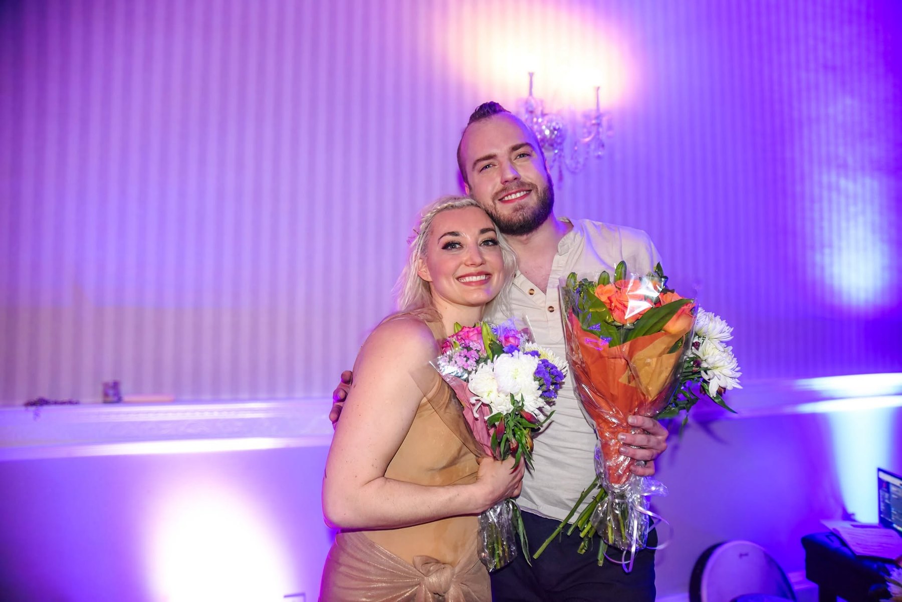 Valev Laube on stage with a performer, both holding bouquets, under purple concert lighting