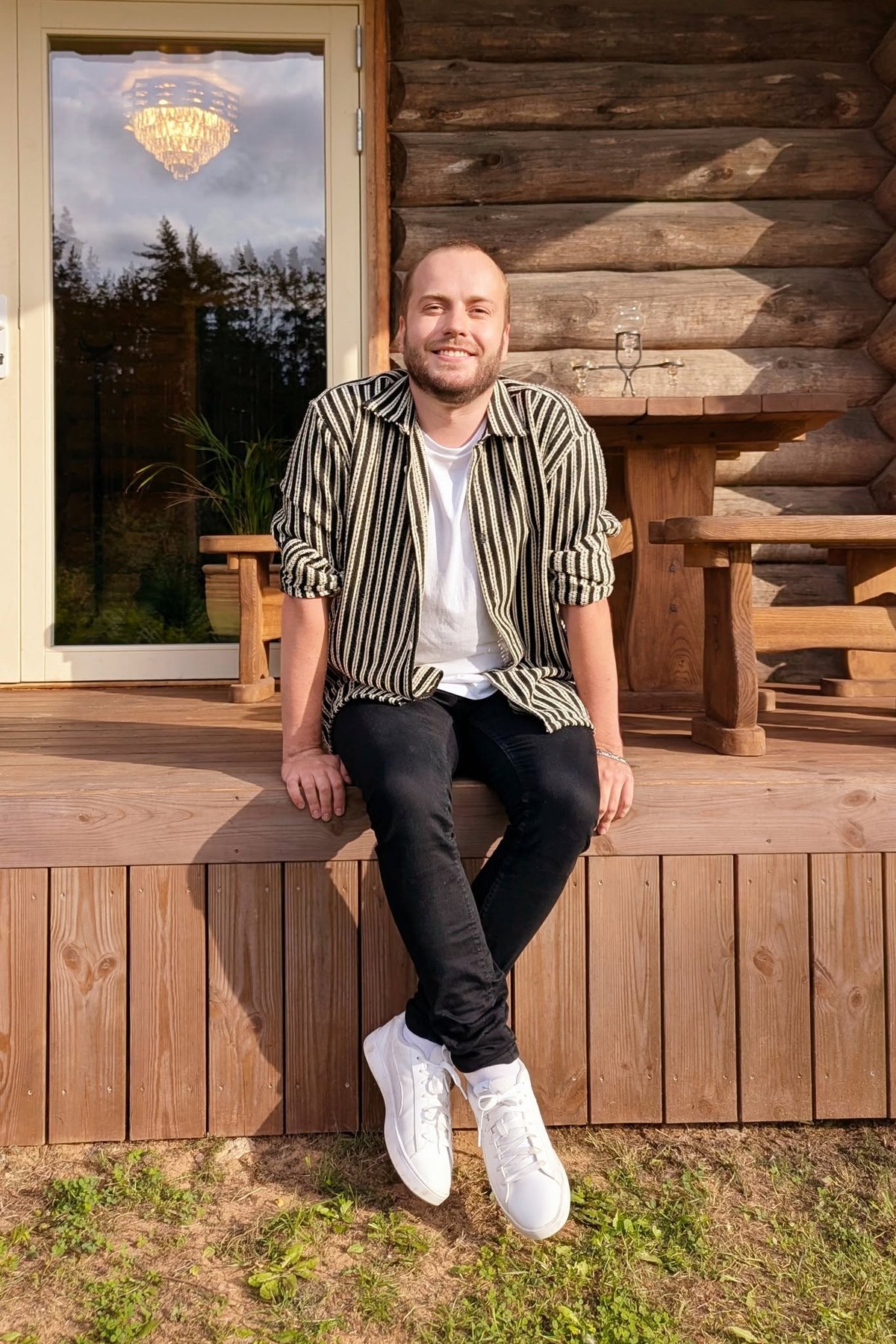 Valev Laube seated on the porch of a log cabin in evening light, in a black and white striped shirt