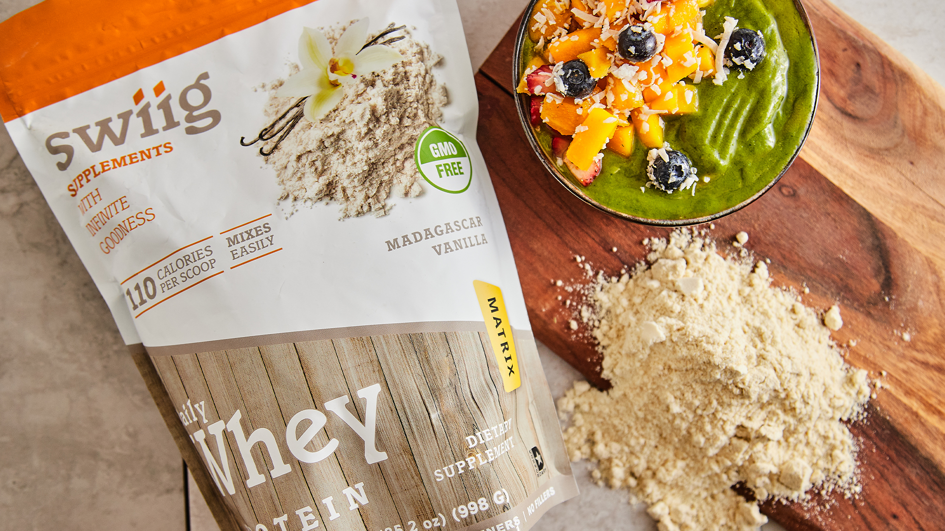 swiig Vanilla Daily Whey Matrix Protein on a kitchen counter next to a wood cutting board and bowl of yogurt and fruit