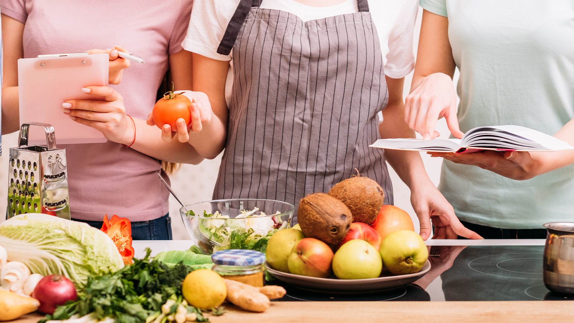 three women at a wooden counter with produce on the counter with notebooks talking about nutrition