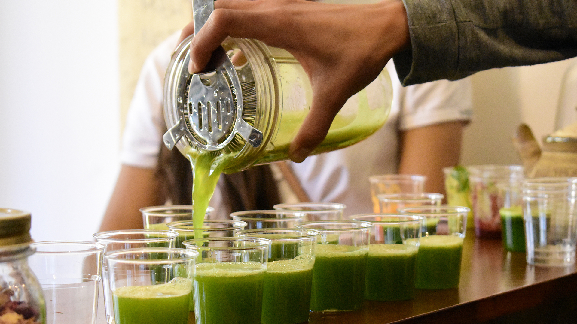 Employee behind smoothie bar counter pouring green protein shake samples into sample cups