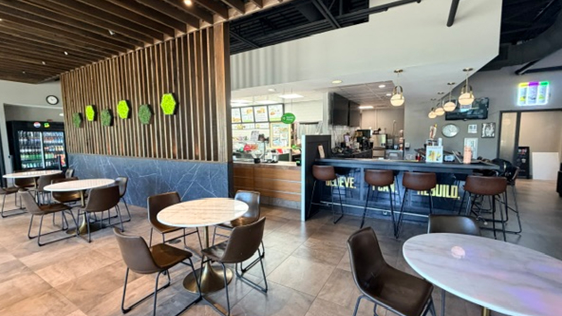 Gym Juice bar with grey marble flooring, wooden accents, grey marble tables with black chairs and a marble countertop with colorful performance food centers smoothie menus behind it to order from
