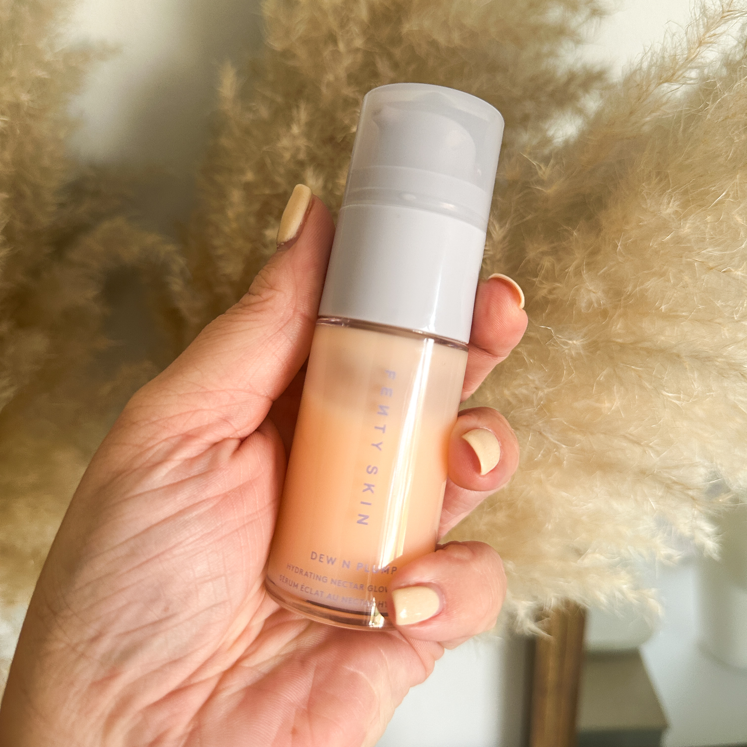 Hand with nude coloured nail polish holding the Fenty Dew N Plump serum in front of pampus grass