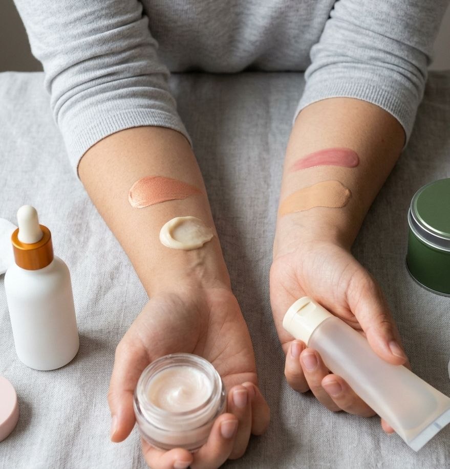 Person swatching makeup and skincare products on forearms, holding a jar and tube against neutral linen background
