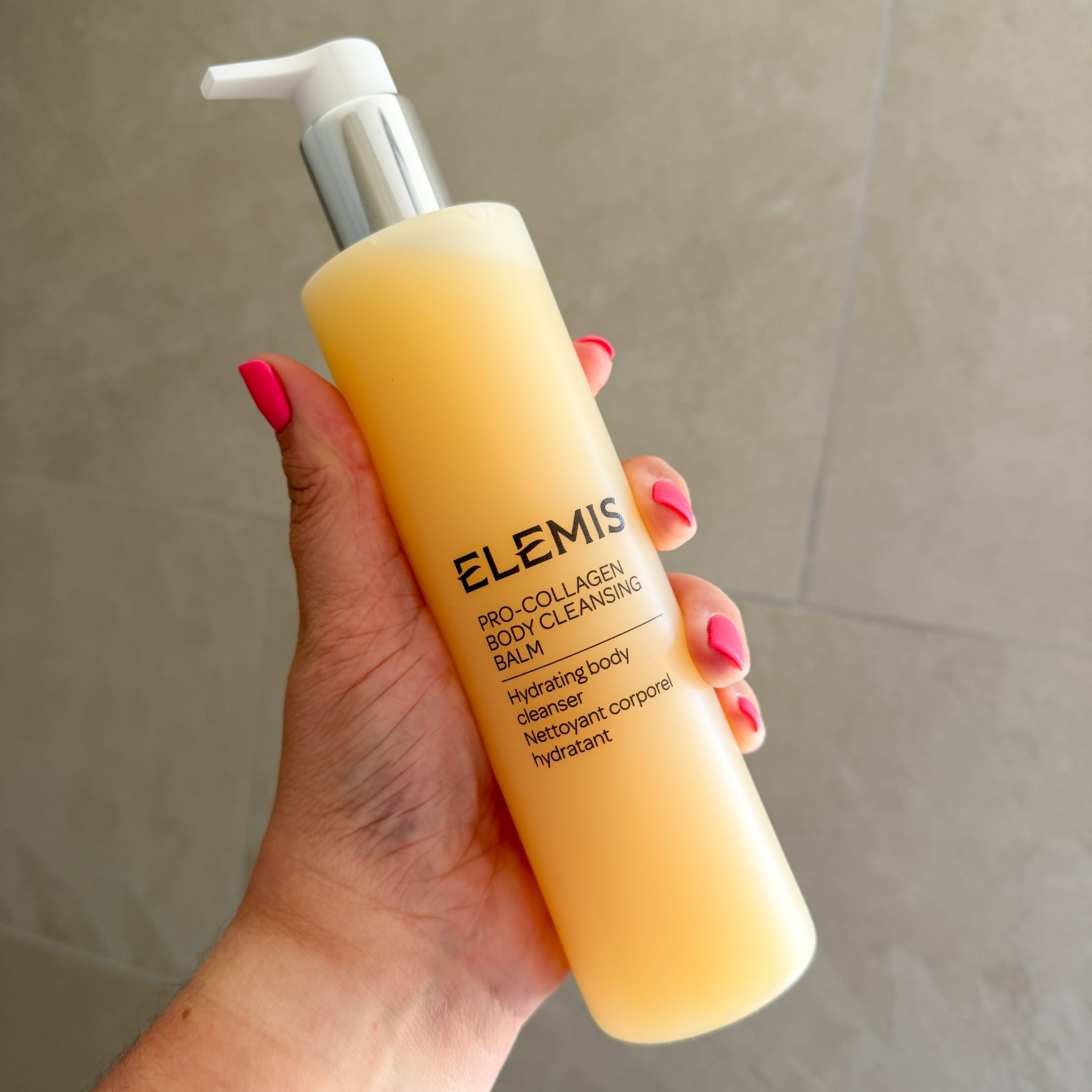 A white hand with neon pink nails holding up the Elemis Body Cleansing Balm against beige bathroom tiles