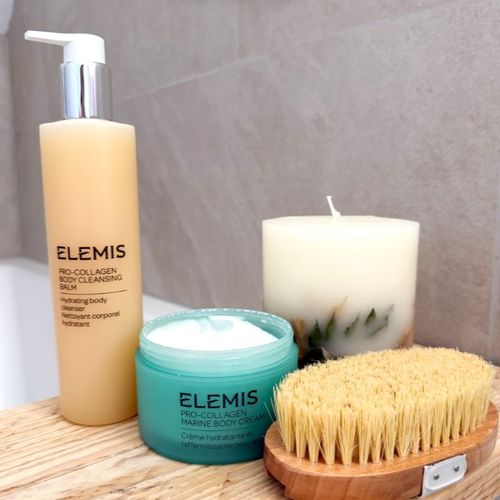 Elemis Pro-Collagen body cleansing balm and marine body cream displayed with a body brush and candle
