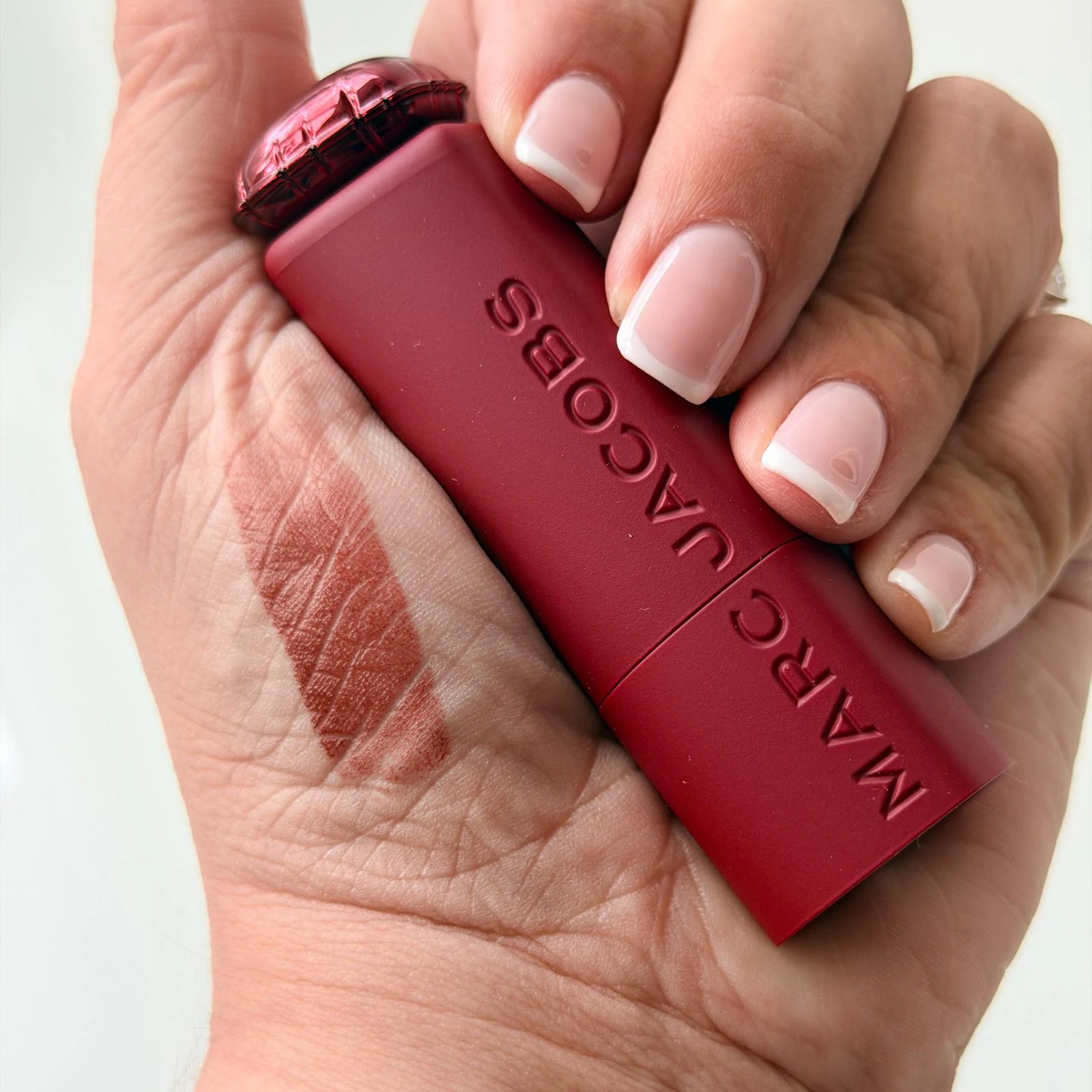 White skin hand holding Marc Jacobs lipstick burgundy packaging with swatch of red shade on thumb pad, nails have French tips