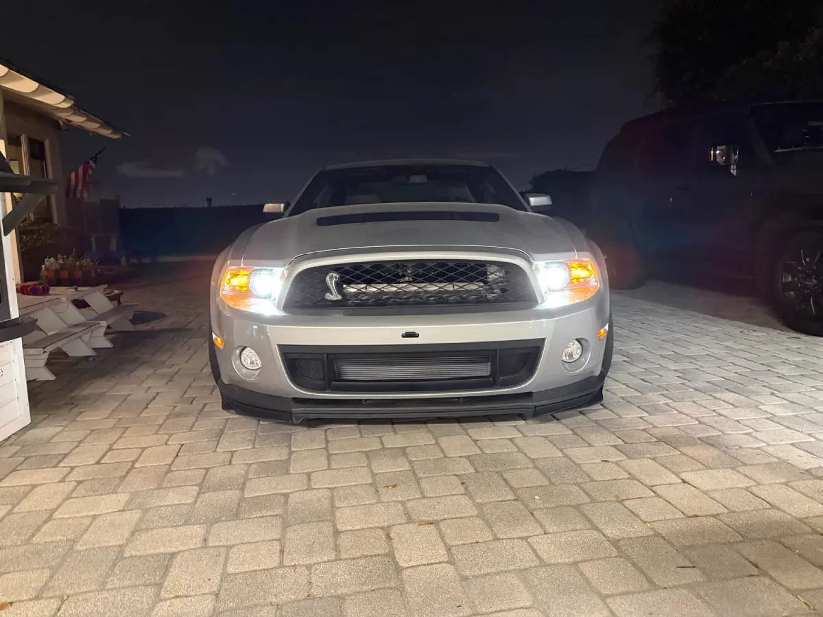 Silver Shelby GT500 in the driveway at night with its headlights on and the heat exchanger visible through the lower grille.