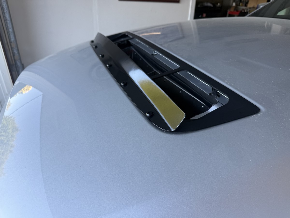 Satin-black aluminum gurney flap bolted along the front of the GT500 hood vent, with the outlet duct below.