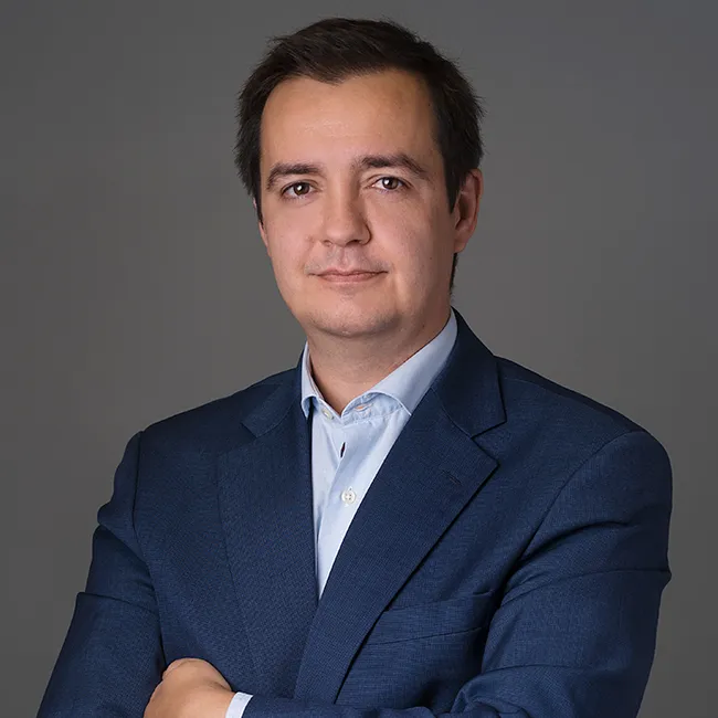 Man with dark hair wearing a navy blue suit jacket and light blue shirt against a gray background.