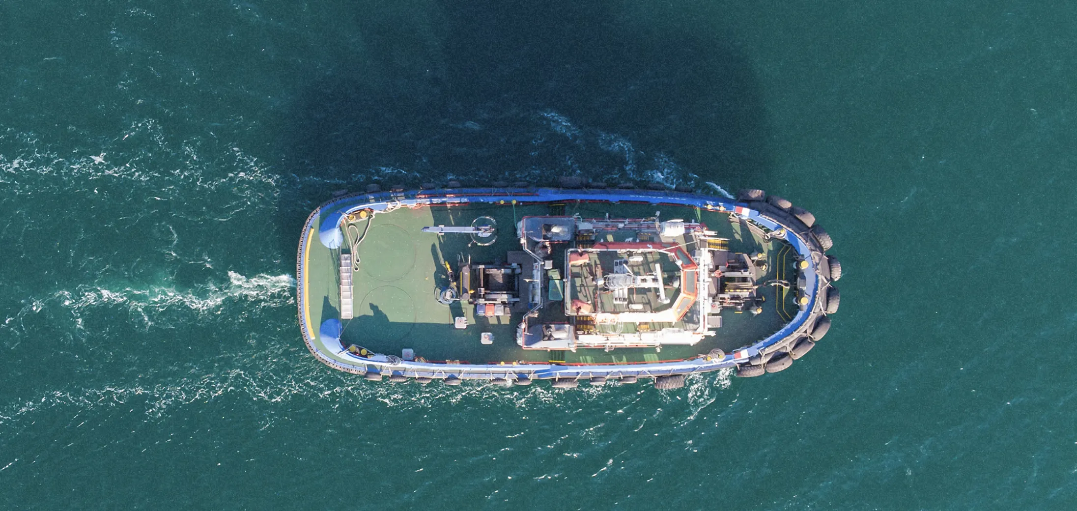 Aerial view of a tugboat sailing on calm blue-green water with visible wake behind it.