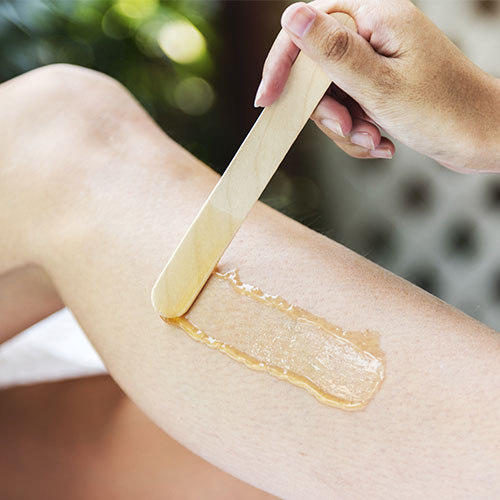 Close-up of a person's hand applying a strip of warm wax on their leg with a wooden spatula for hair removal.