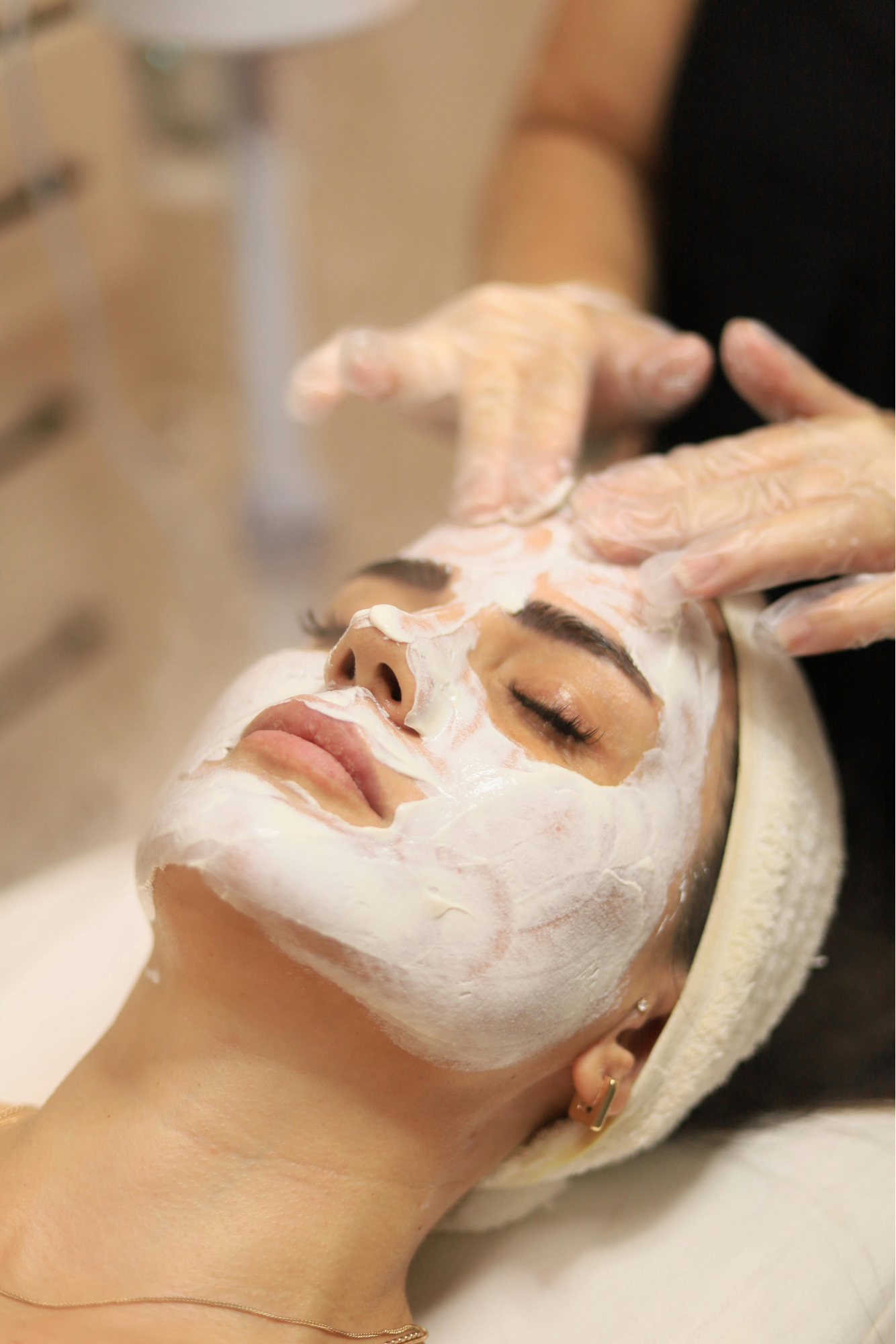 Close-up of a woman lying down with eyes closed, receiving a facial treatment with white cream mask being applied by gloved hands, wearing a headband.