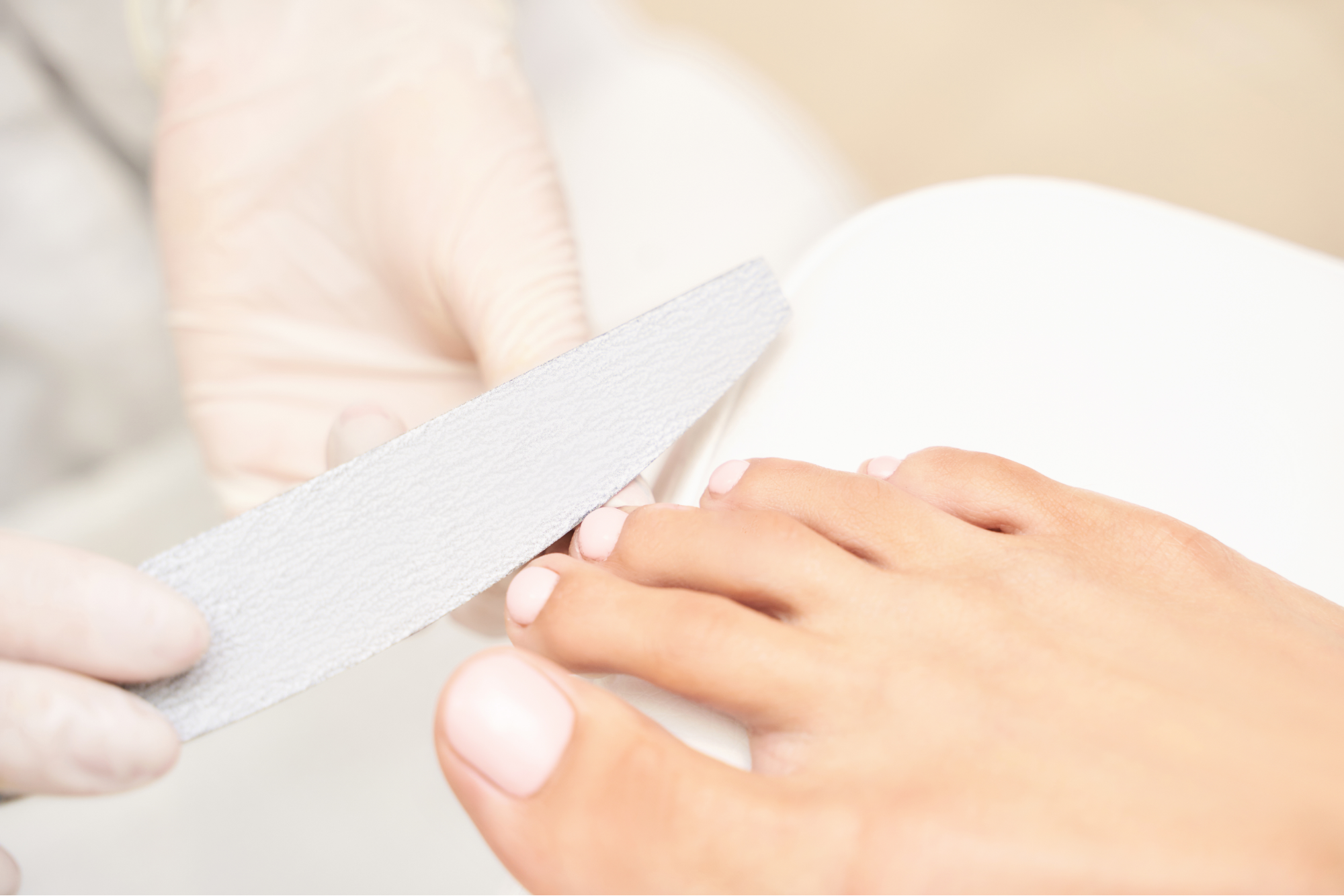Close-up of a person filing toenails with a nail file held by hands wearing disposable gloves, on feet with light pink nail polish.