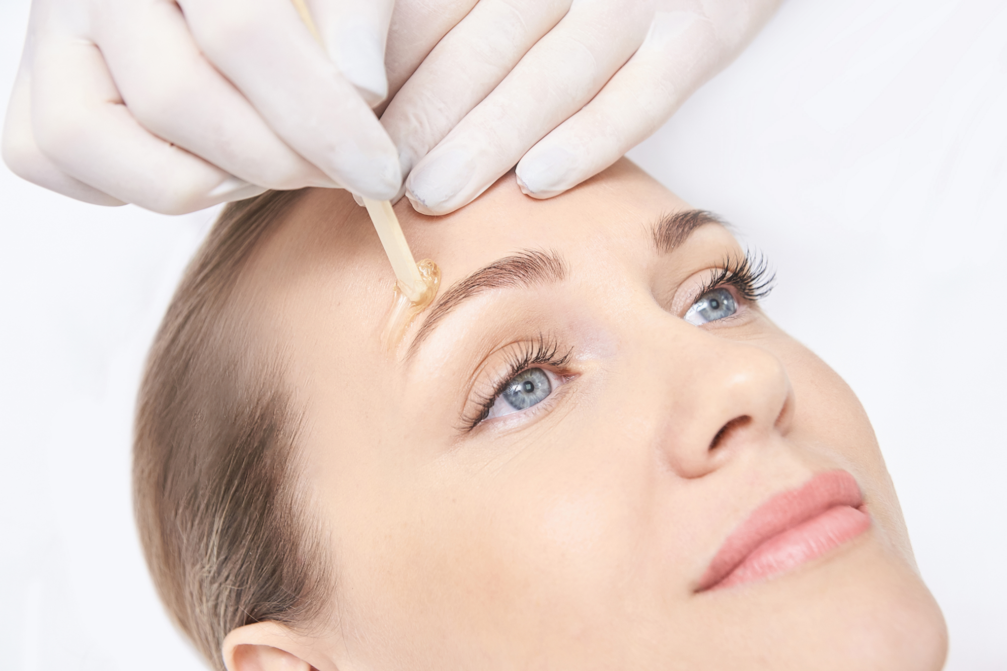 Close-up of a woman with blue eyes receiving eyebrow waxing; gloved hands apply warm wax with a wooden stick to her brow.