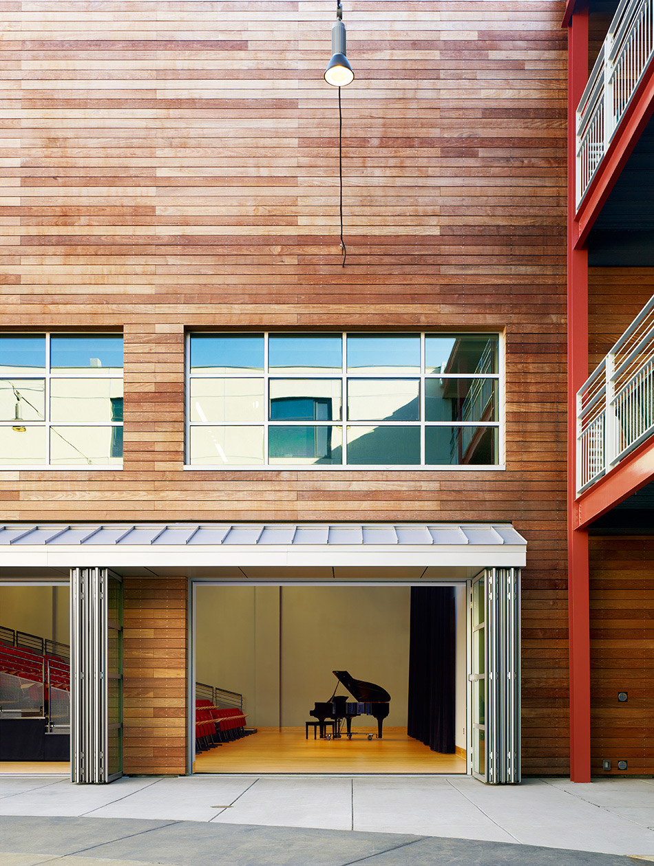 Drew School New Assembly Wing, San Francisco, CA: Education new construction by Herrero Builders with LEED Gold sustainable design, with ROMA Design Group. Exterior view of the wood-clad wall with an open folding glass door revealing a grand piano on stage.