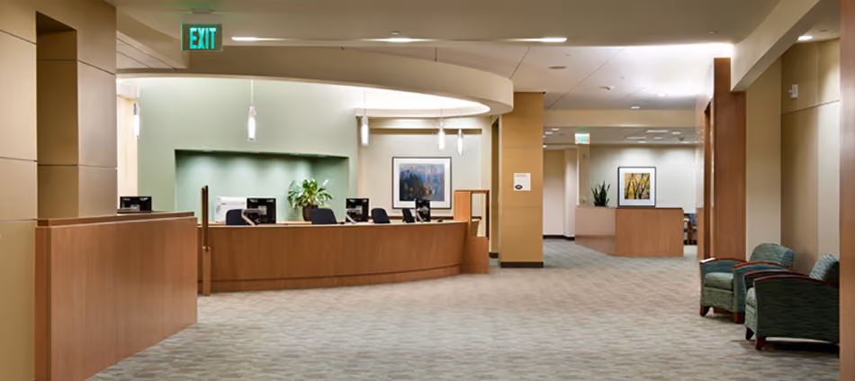 Sutter Health PAMF Burlingame Medical Office Building, Burlingame, CA: Healthcare Tenant Improvements by Herrero Builders under Integrated Project Delivery, with Hawley Peterson Snyder. Interior view of a curved wood reception desk under a soffit, with green seating and a long corridor beyond.
