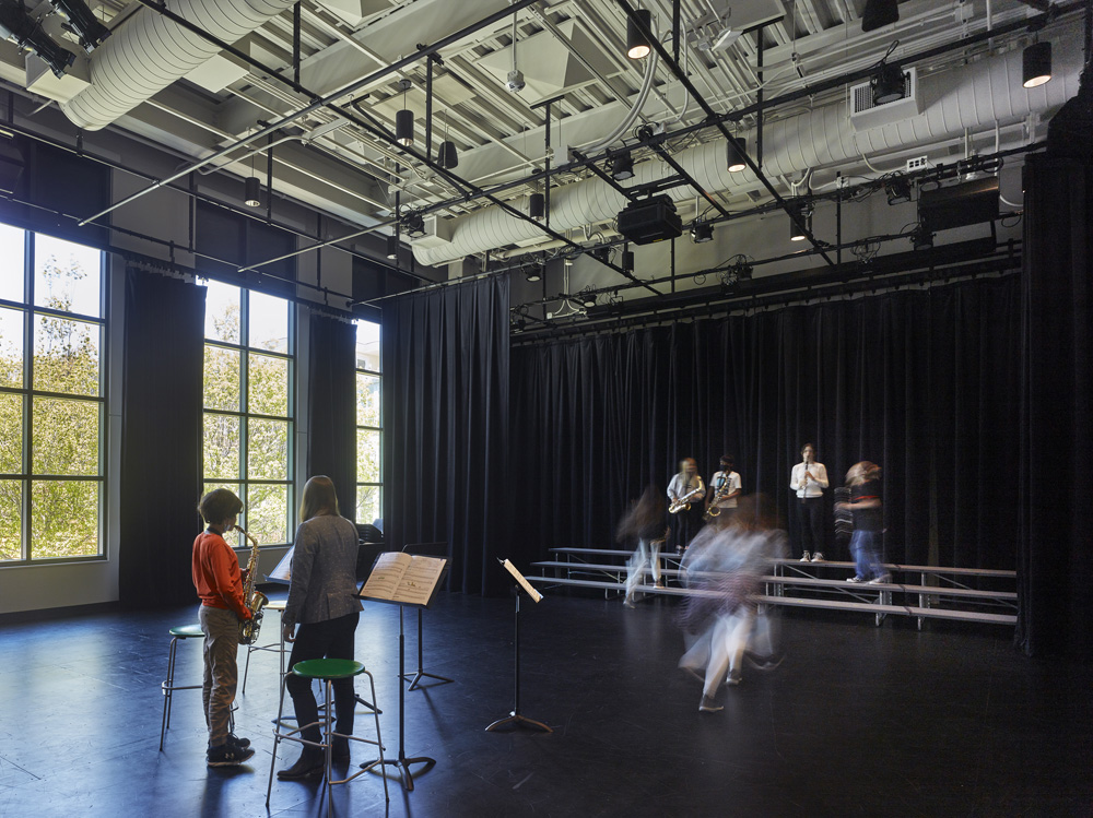 San Francisco Day School School Addition and Staff Housing, San Francisco, CA: Education new construction by Herrero Builders under Integrated Project Delivery, with Studio Bondy. Interior view of a dark performance room with exposed ceiling ducts, tall windows and students rehearsing on risers.