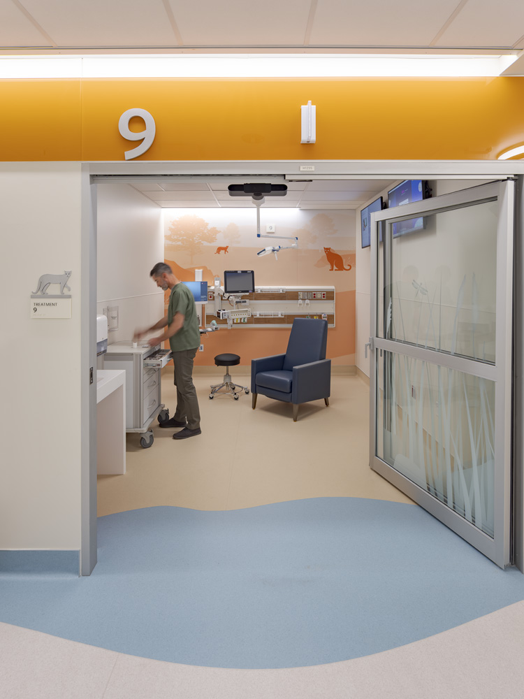 Stanford Health Care Stanford Health Pediatric Emergency Department, Palo Alto, CA: Healthcare renovation by Herrero Builders in an occupied hospital, with Ratcliff. Interior view of an exam room entrance with an orange header, number 9, sliding glass door and blue floor curve.