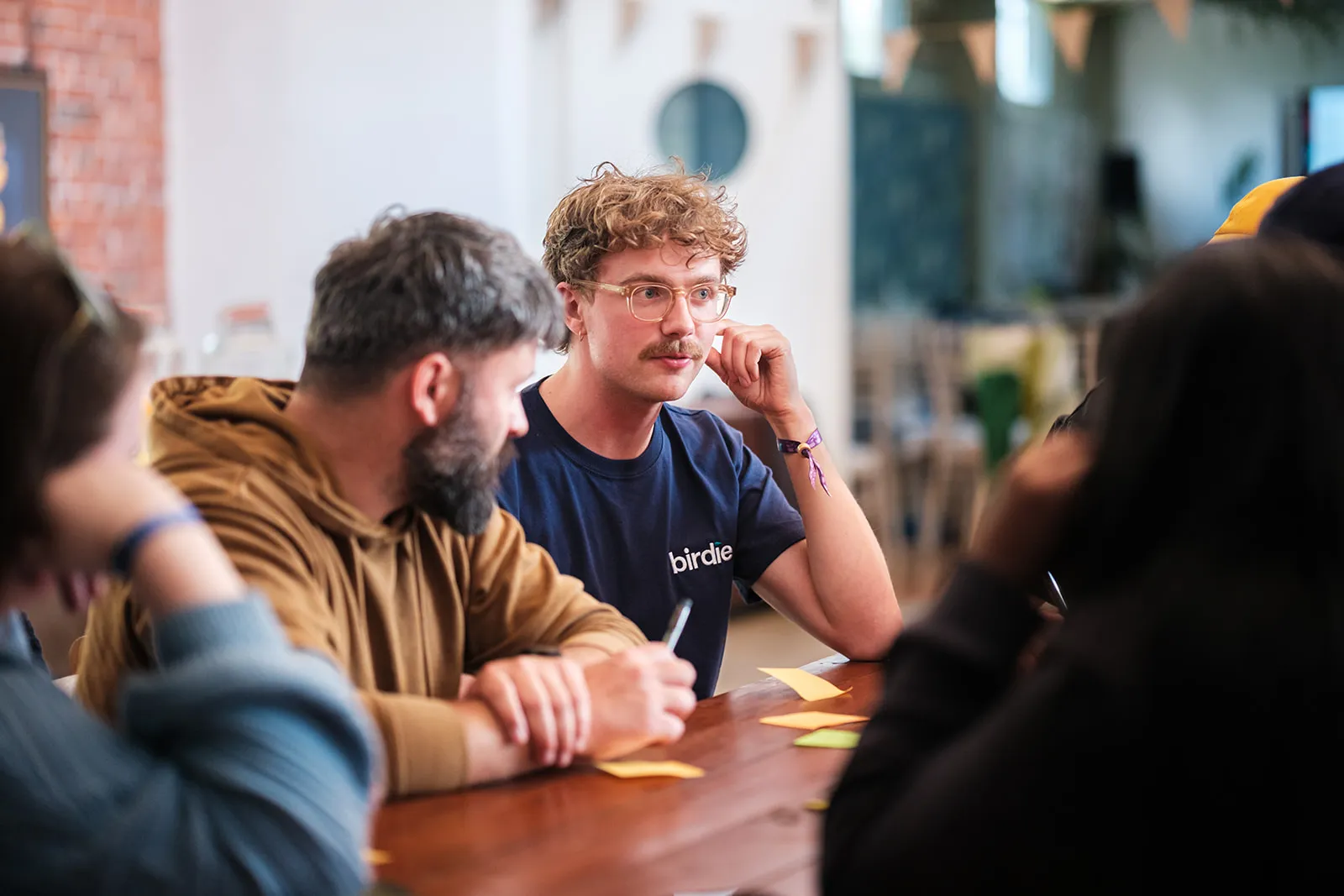 Young man with glasses and mustache listens attentively during a group meeting at a wooden table.