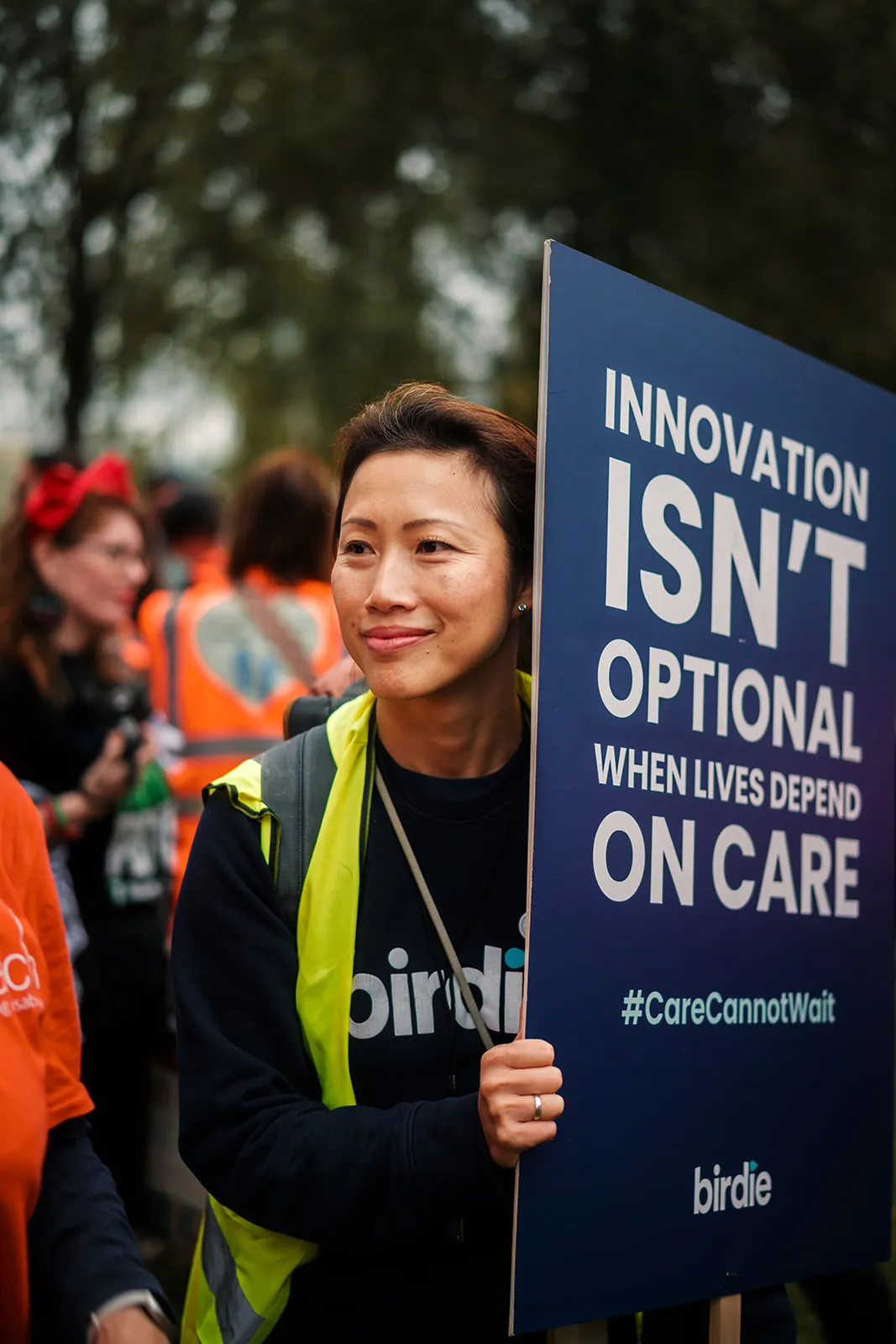 Person wearing yellow vest holding sign saying innovation isn't optional for care with hashtag CareCannotWait.
