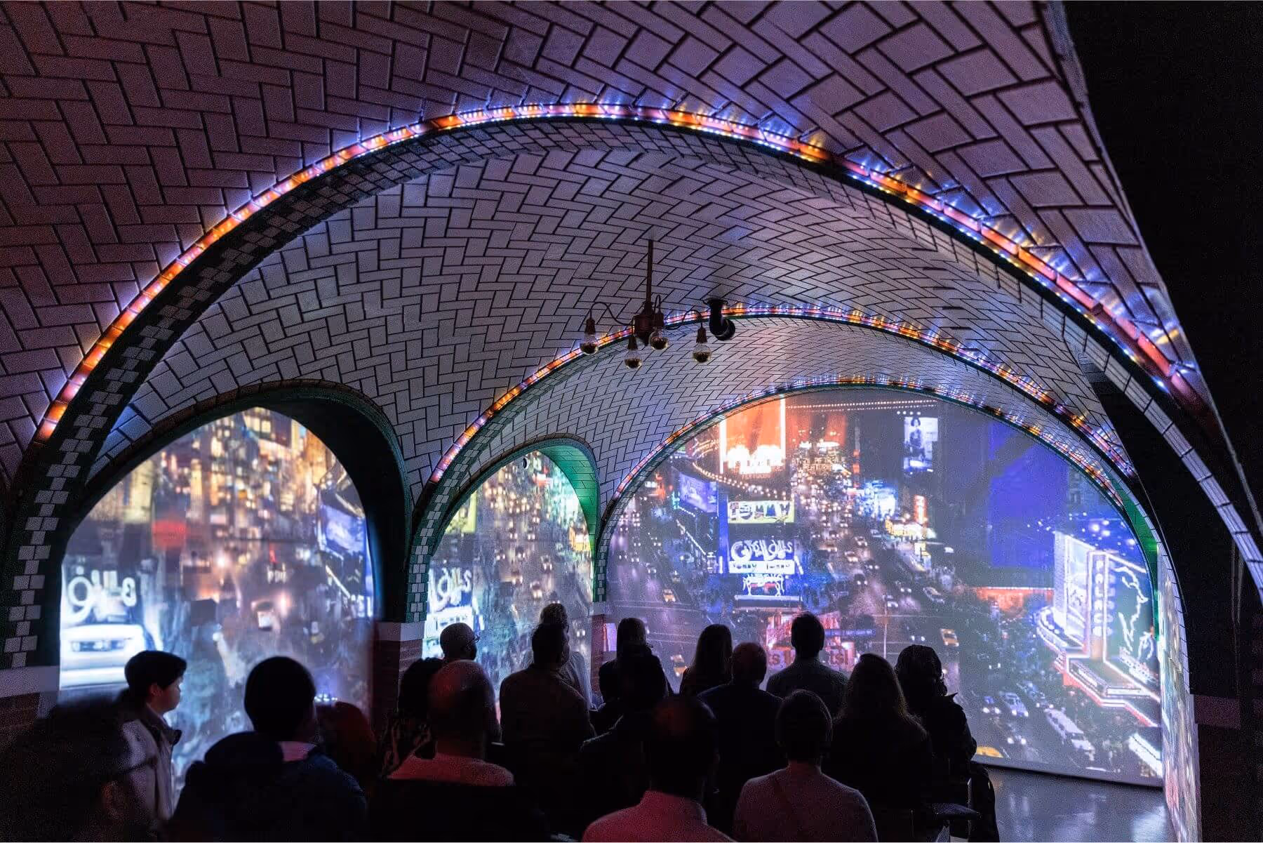 A section of RiseNY, showcasing multiple screens in a room with people sitting on benches and watching the screens.