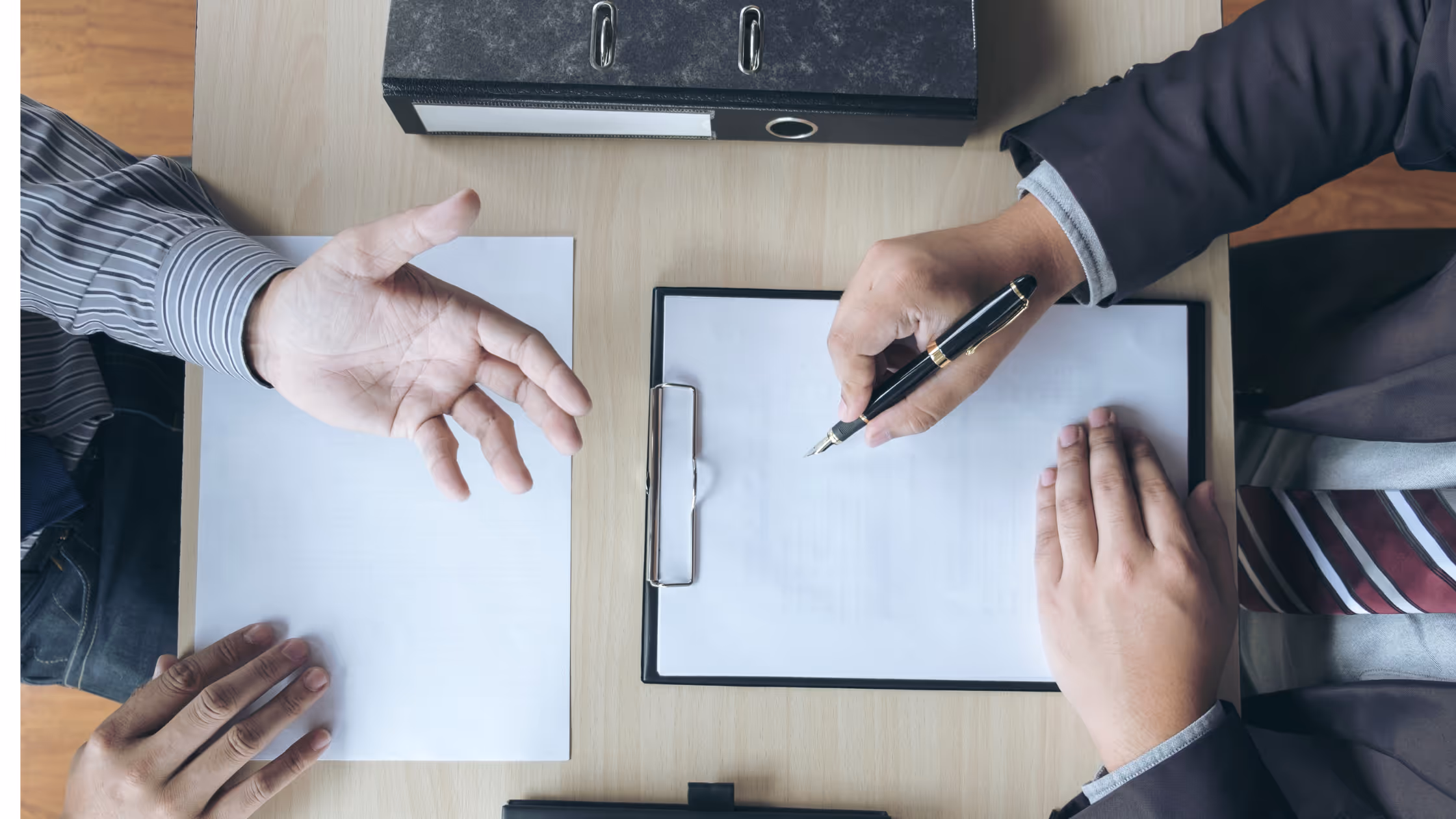 Man in a suit writes notes on a clipboard while wearing a suit. Man opposite gestures with his hands.