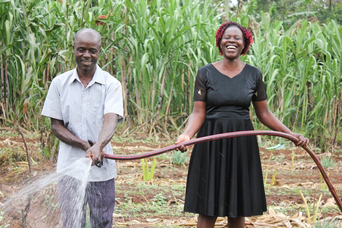 A man and woman stand in a field watering crops with a hose, both smiling with tall green plants behind them.