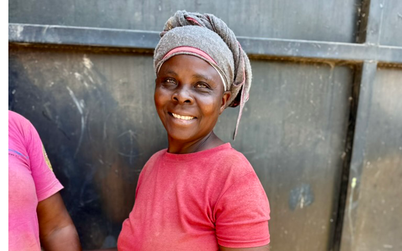 A woman stands smiling in front of a dark metal wall, wearing a red shirt, headscarf, and work gloves, with another person partly visible beside her.