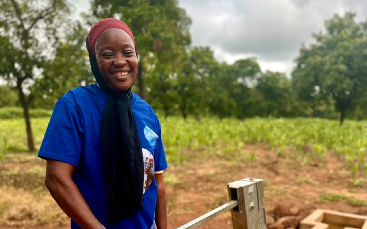 A woman smiles while pumping water from a hand pump in an outdoor rural setting, with fields and trees behind her.