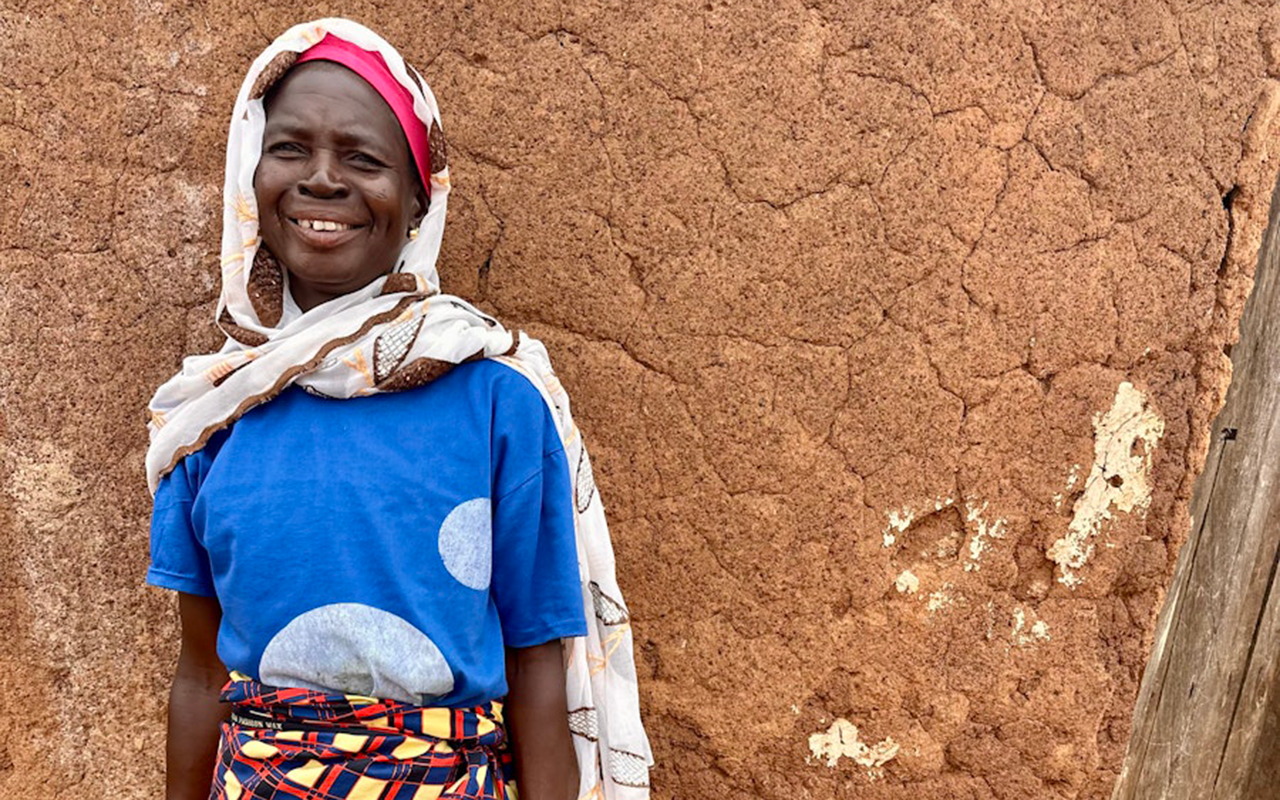 A woman stands smiling against an earthen wall, dressed in a blue shirt, patterned wrap skirt, and headscarf.A woman stands smiling against an earthen wall, dressed in a blue shirt, patterned wrap skirt, and headscarf.