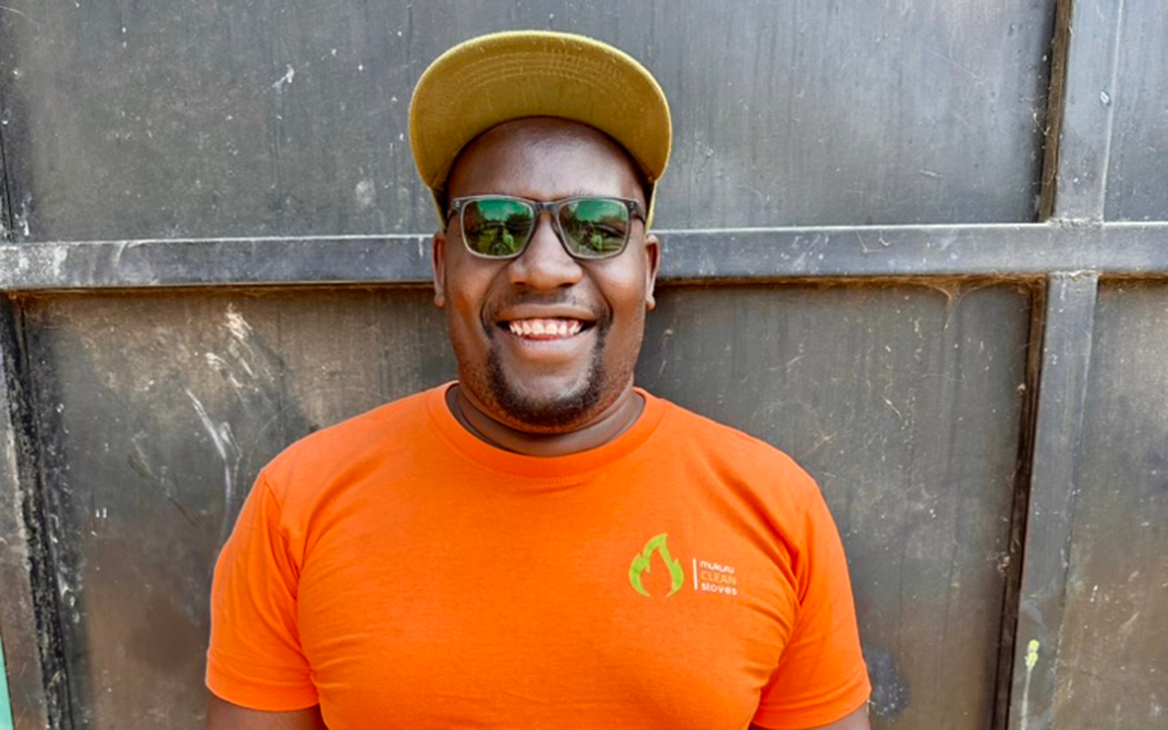 A man stands smiling in front of a dark metal wall, wearing sunglasses, an orange T-shirt, and a yellow cap.