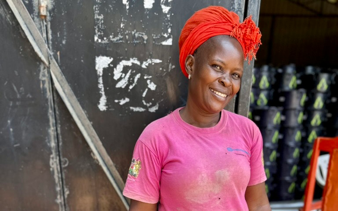 A woman stands smiling in front of a dark metal doorway, wearing a bright headwrap, pink shirt, and patterned skirt, with her hands dusted from work.