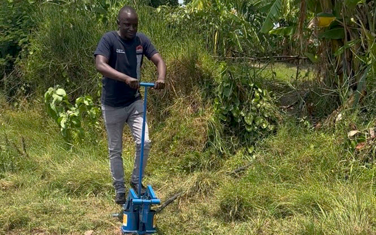 A man operates a blue manual water pump in a grassy farm field, with banana trees and lush vegetation in the background.