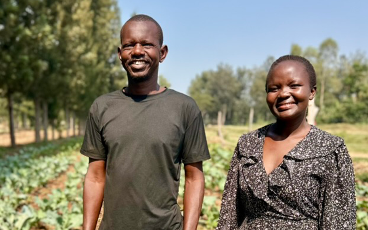 A man and woman stand smiling together in a field of leafy crops, with tall trees and a clear blue sky in the background.