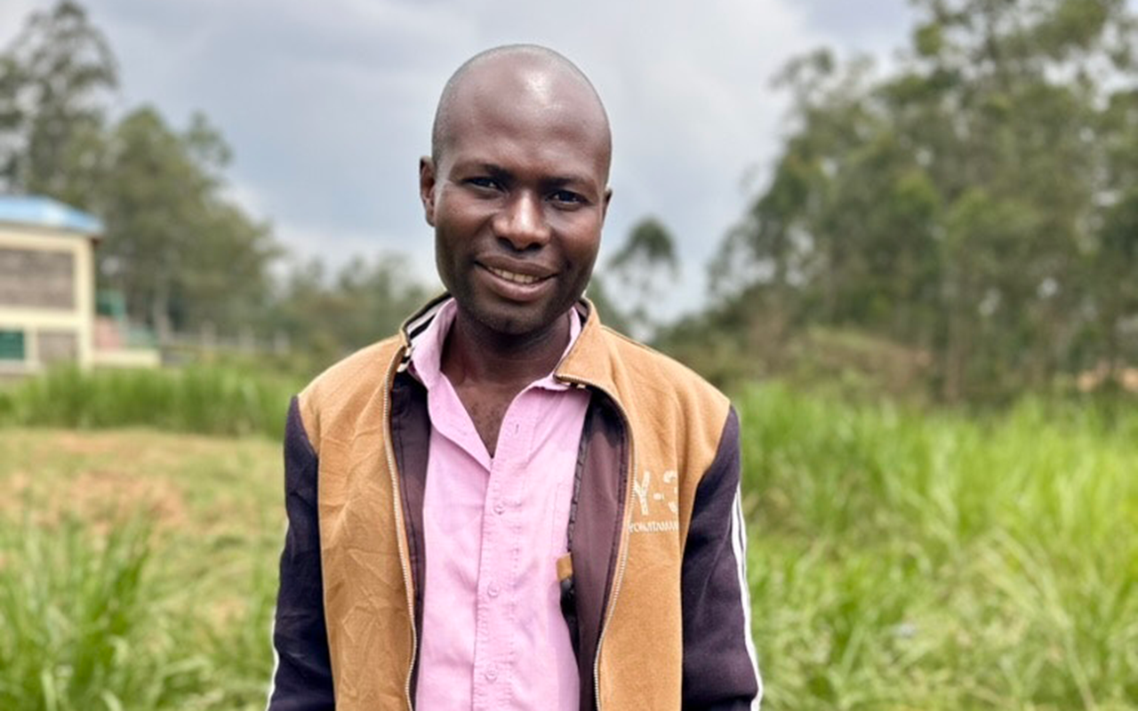 A man stands smiling in a field with small leafy crops, wearing a light pink shirt and tan-and-black jacket, with trees and sky in the background.