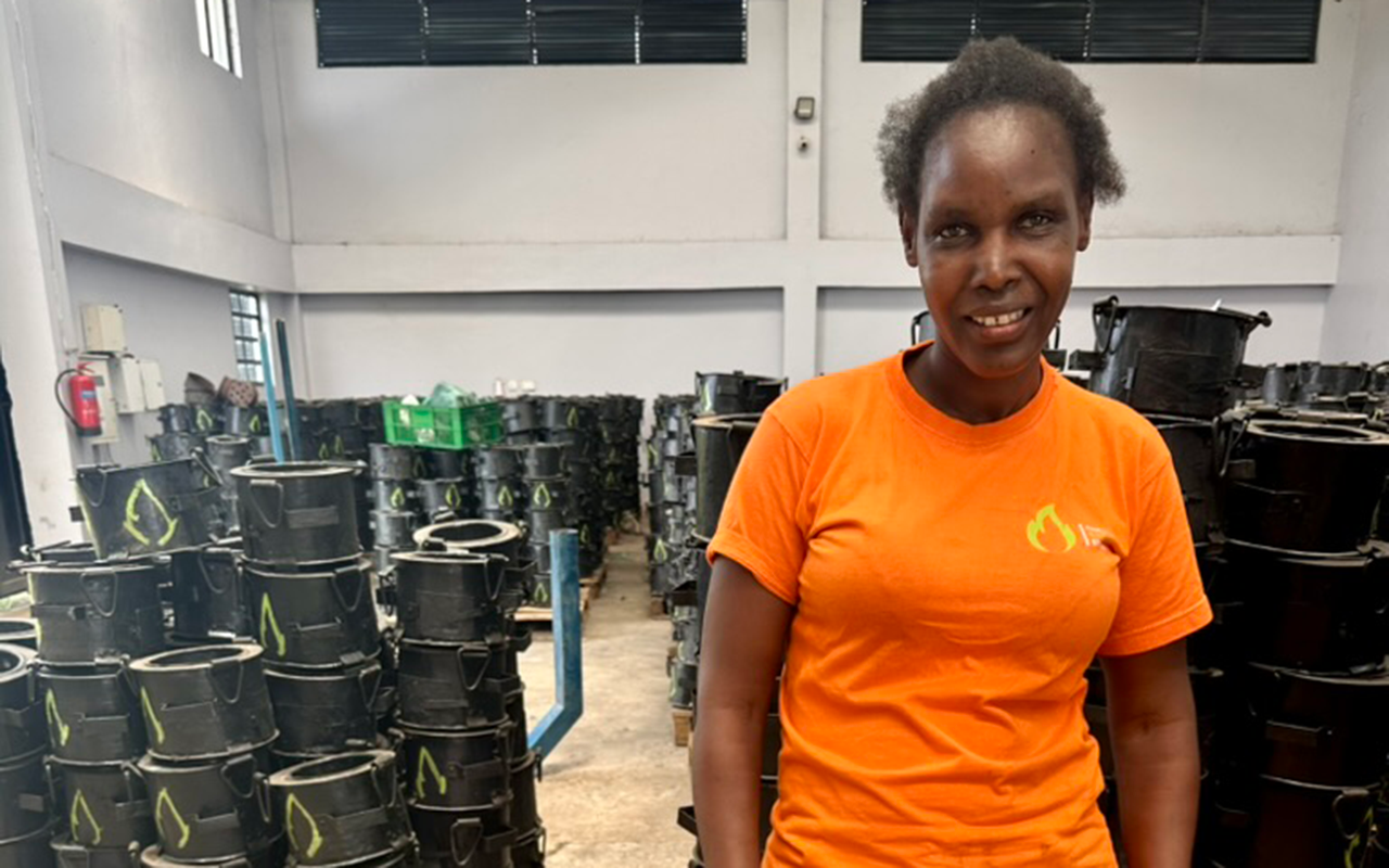A woman stands smiling inside a workshop, wearing an orange shirt, surrounded by stacks of black clean-cookstove parts.