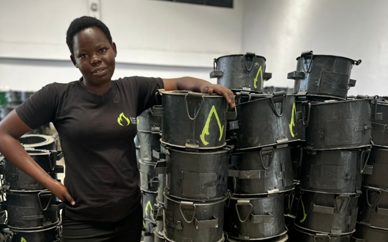 A woman stands indoors beside tall stacks of black clean-cookstoves, resting her arm on them and looking toward the camera.