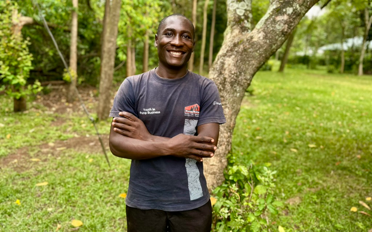 A man stands outdoors in a grassy, wooded area with his arms crossed, smiling toward the camera.