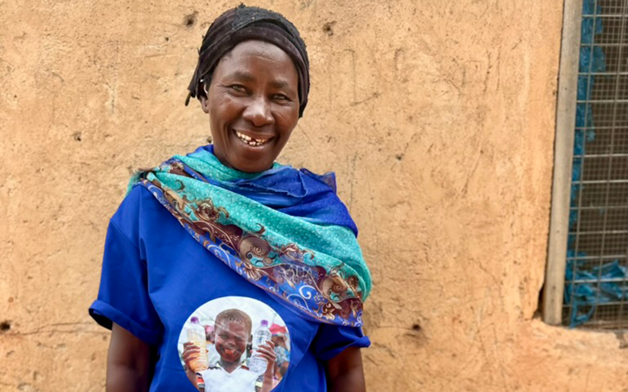 A woman stands smiling in front of an earthen wall, wearing a blue shirt with a printed photo on it and a colorful scarf draped over her shoulders.