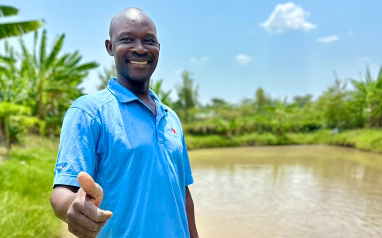 A man stands by a small pond on a sunny day, smiling and giving a thumbs-up, with green plants and trees in the background.
