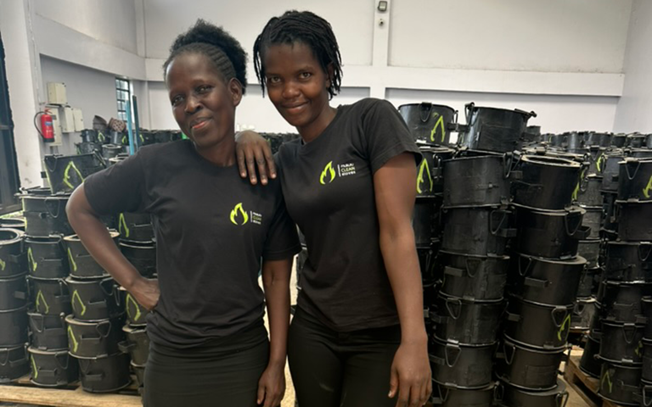 Two women stand together smiling inside a workshop, wearing black shirts, surrounded by tall stacks of black clean-cookstoves.