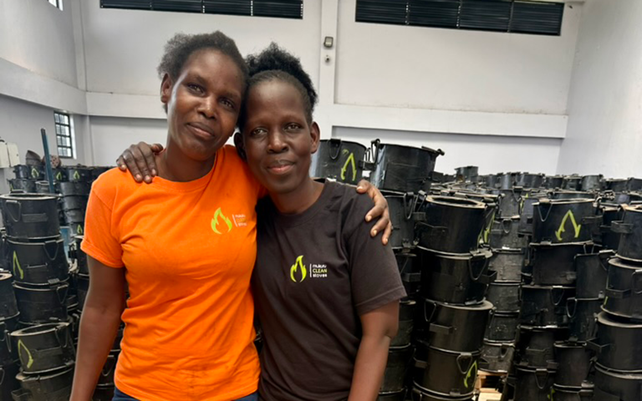 Two women stand close together smiling inside a workshop, one in an orange shirt and one in a black shirt, surrounded by stacks of black clean-cookstoves.