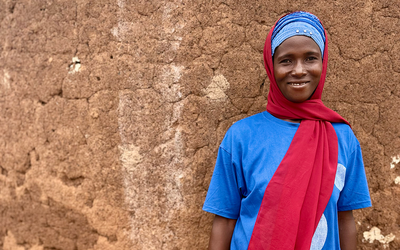 A woman stands smiling against an earthen wall, wearing a blue shirt and a red scarf draped over her shoulder and head.