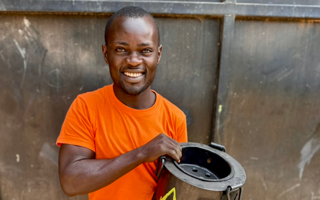 A man stands smiling in front of a dark metal wall, wearing an orange shirt and holding a black clean-cookstove
