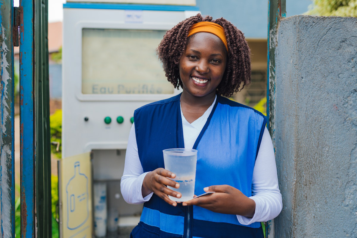 A woman holds a cup of clean water while standing next to a water purification system in Rwanda.